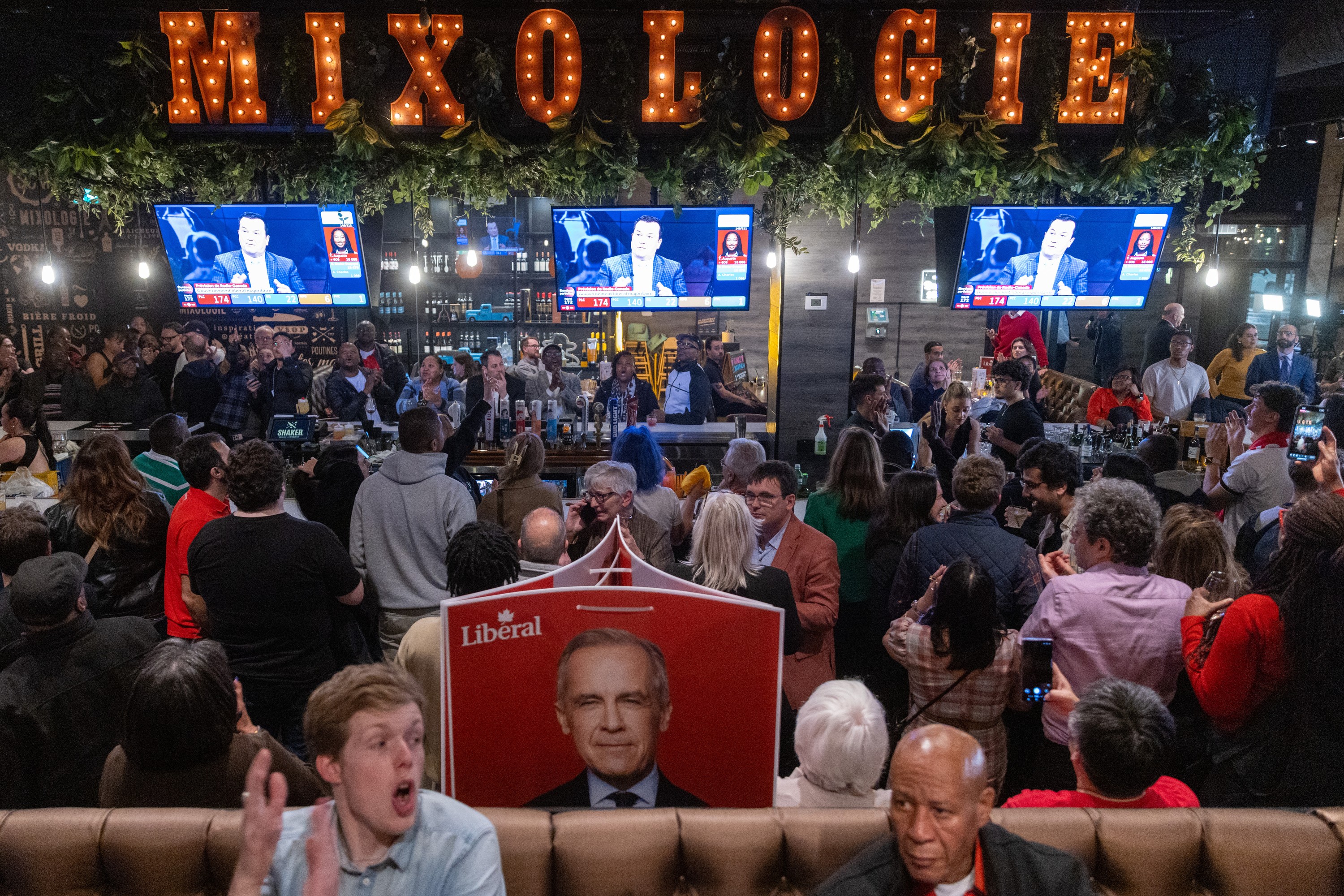 People gather at a bar, a campaign poster with Mark Carney's face is visible.