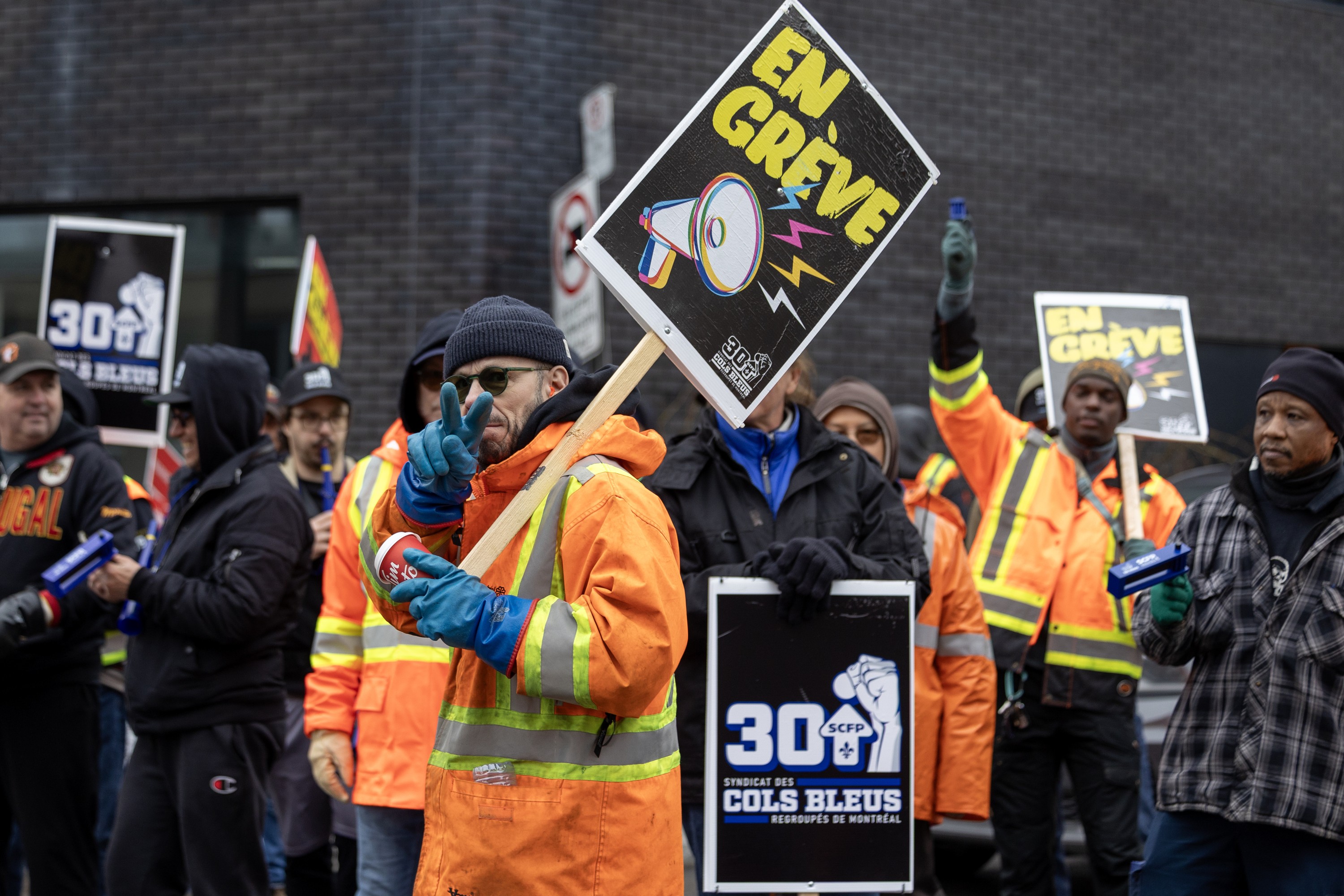 A man holds an on strike sign and holds up two fingers at the camera while picketing.