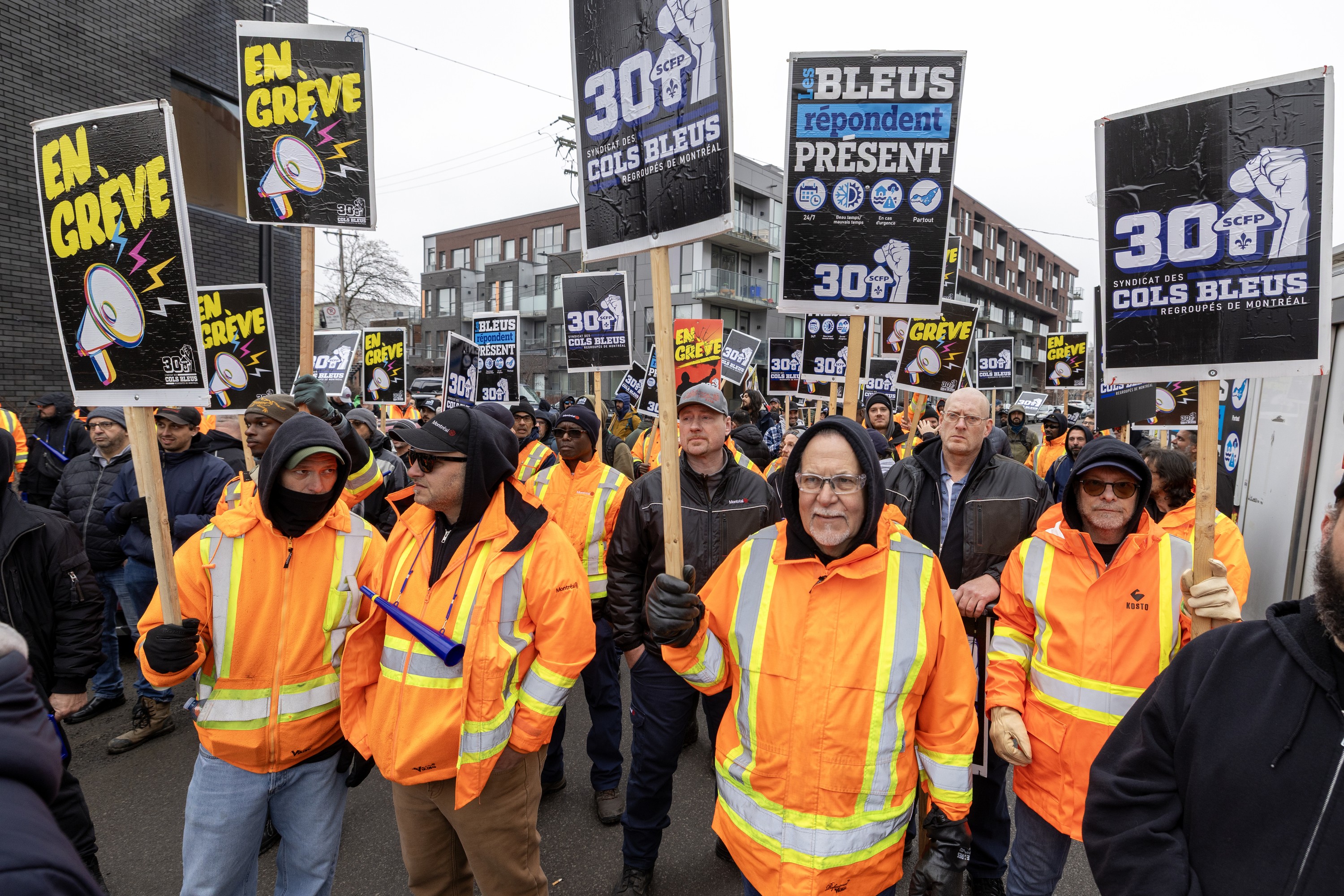 A large group of protesters, some in orange safety vests, protest outside.
