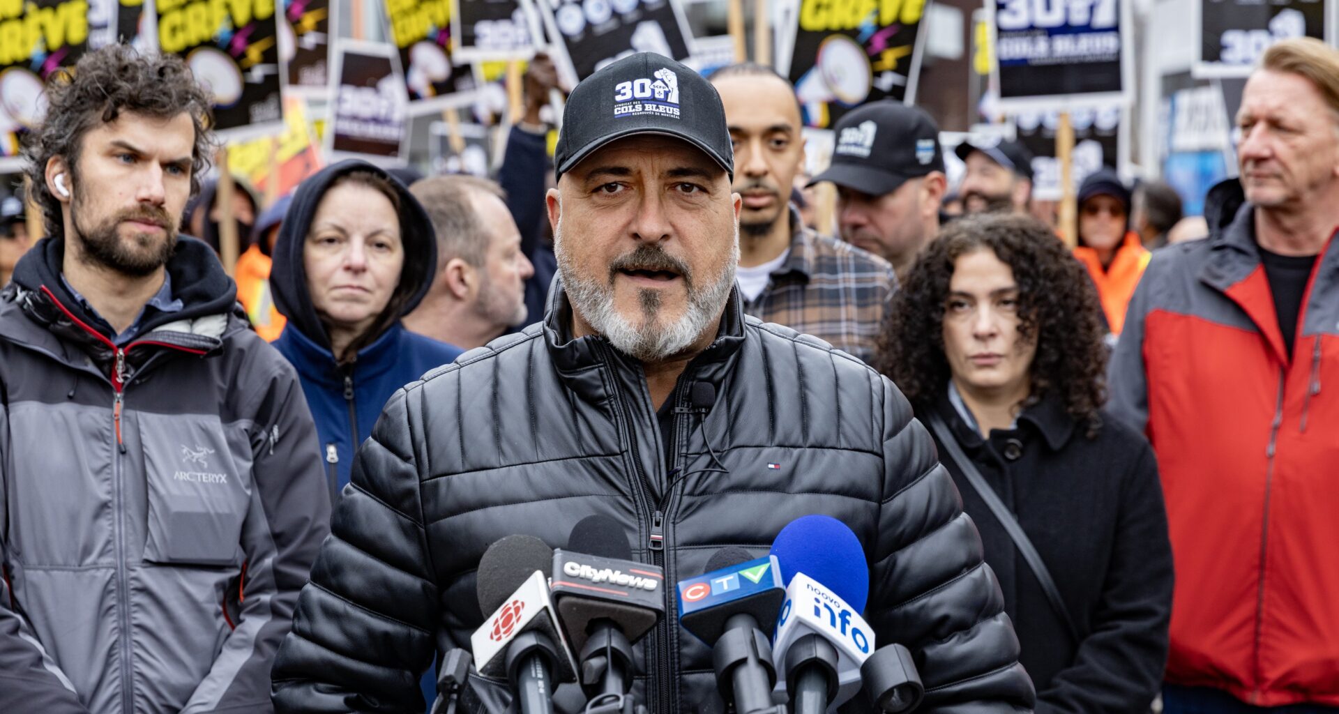 Jean-Pierre Lauzon speaks into several news media microphones while a large group of people, some carrying union signs, stands behind him.