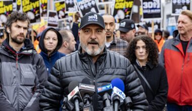 Jean-Pierre Lauzon speaks into several news media microphones while a large group of people, some carrying union signs, stands behind him.