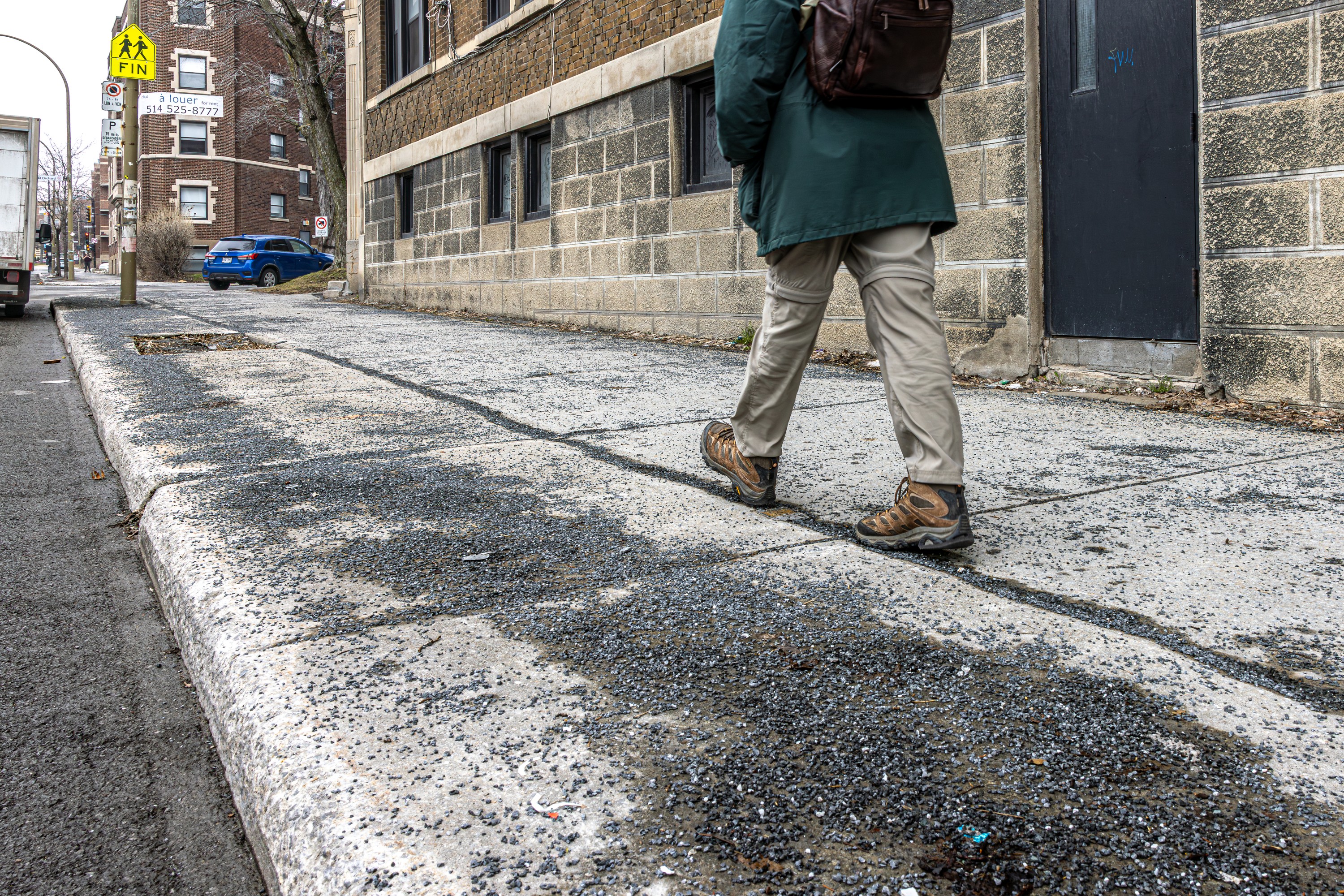 A man walks on a sidewalk covered by gravel from winter. 