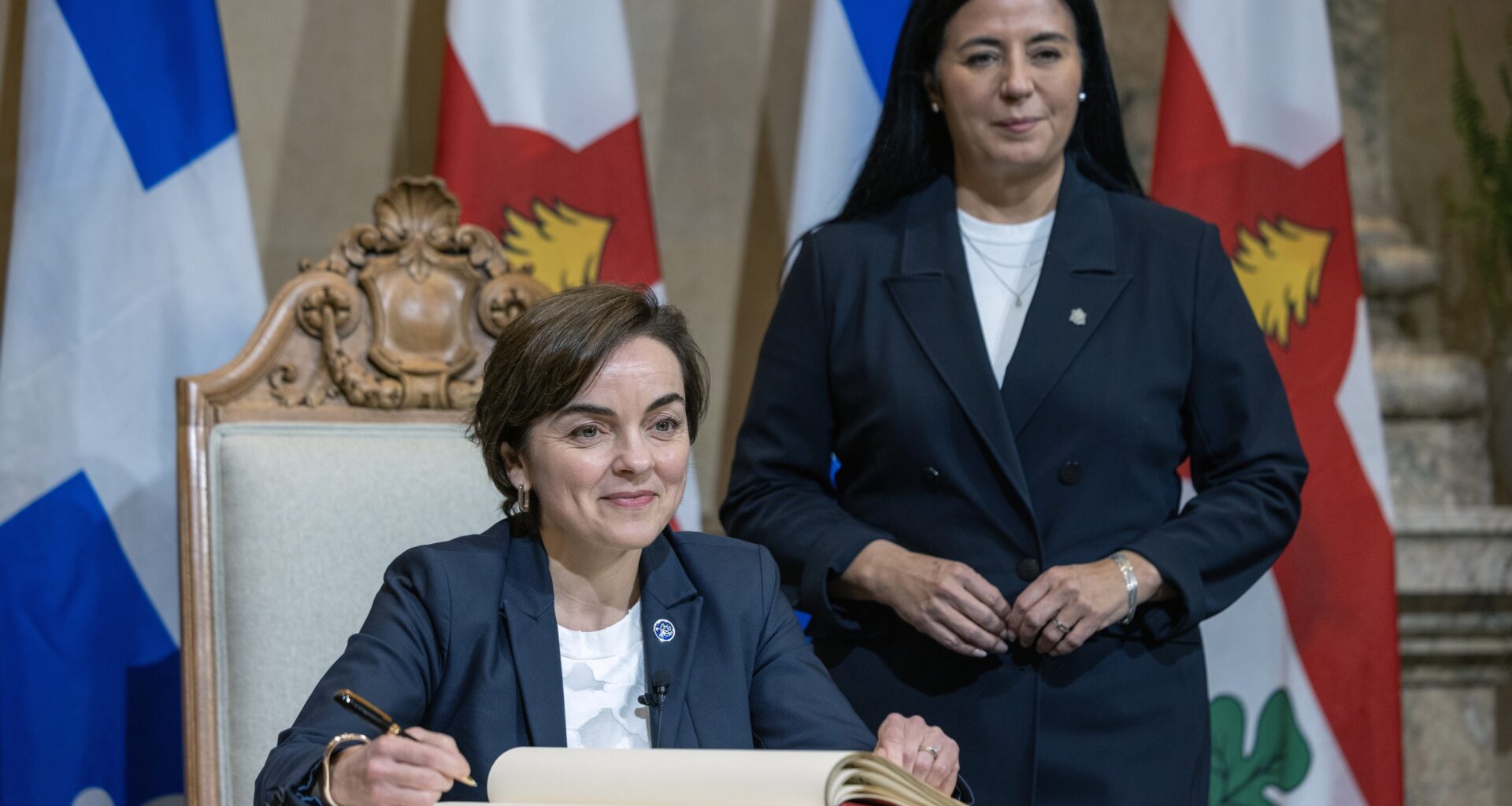 Quebec Premier Christine Fréchette sits while signing a book, with Montreal Mayor Soraya Martinez Ferrada standing behind her.
