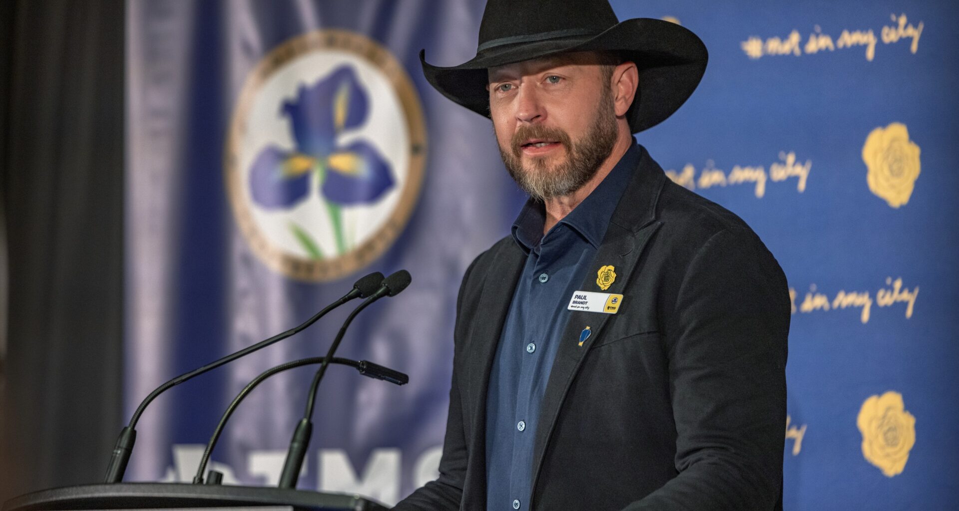 Paul Brandt, wearing a black cowboy hat, stands at a podium with microphones.