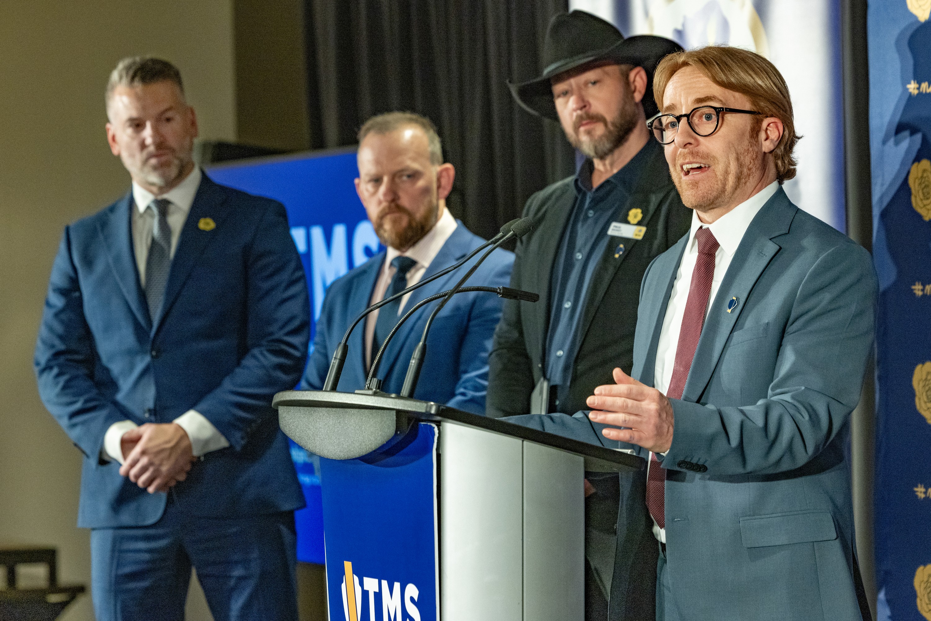 Three men in suits, one wearing a black cowboy hat, listen to a fourth man in a suit speak at a podium.