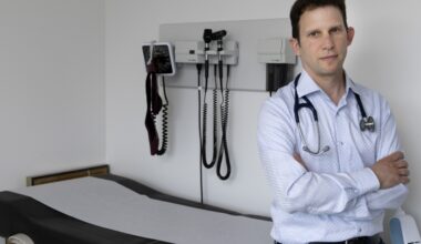 A doctor with his arms crossed, leaning against an examination table.
