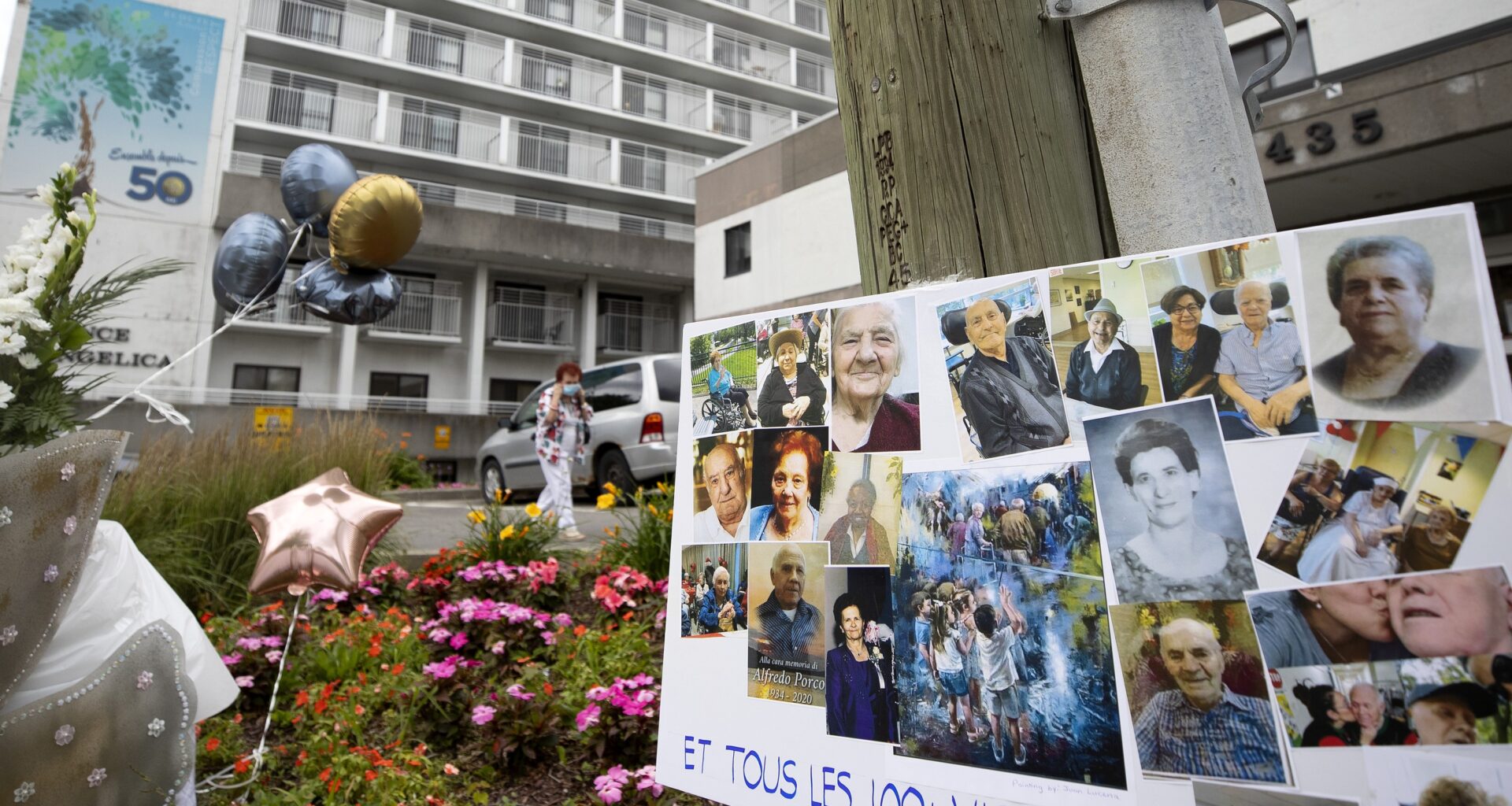 A placard with photographs of family members reads Et tous les 100+ victimes. Behind the placard is a white building, several balloons and a woman putting on a mask.