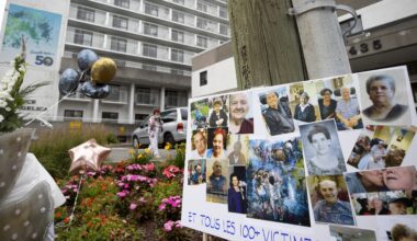 A placard with photographs of family members reads Et tous les 100+ victimes. Behind the placard is a white building, several balloons and a woman putting on a mask.