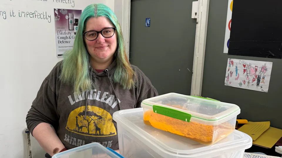 Image shows a women wearing a brown hoodie is smiling at the camera. She has blue shoulder length hair and black rimmed glasses. She is stood behind plastic containers of a yellow spice.