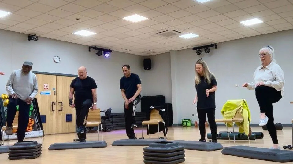 A photo of a group of people doing a workout they are all raising their legs on plastic blocks. There are five people in the photo, two women and three men. None of them is smiling. The rest of the room is very bright with wooden floors and wooden chairs behind them.