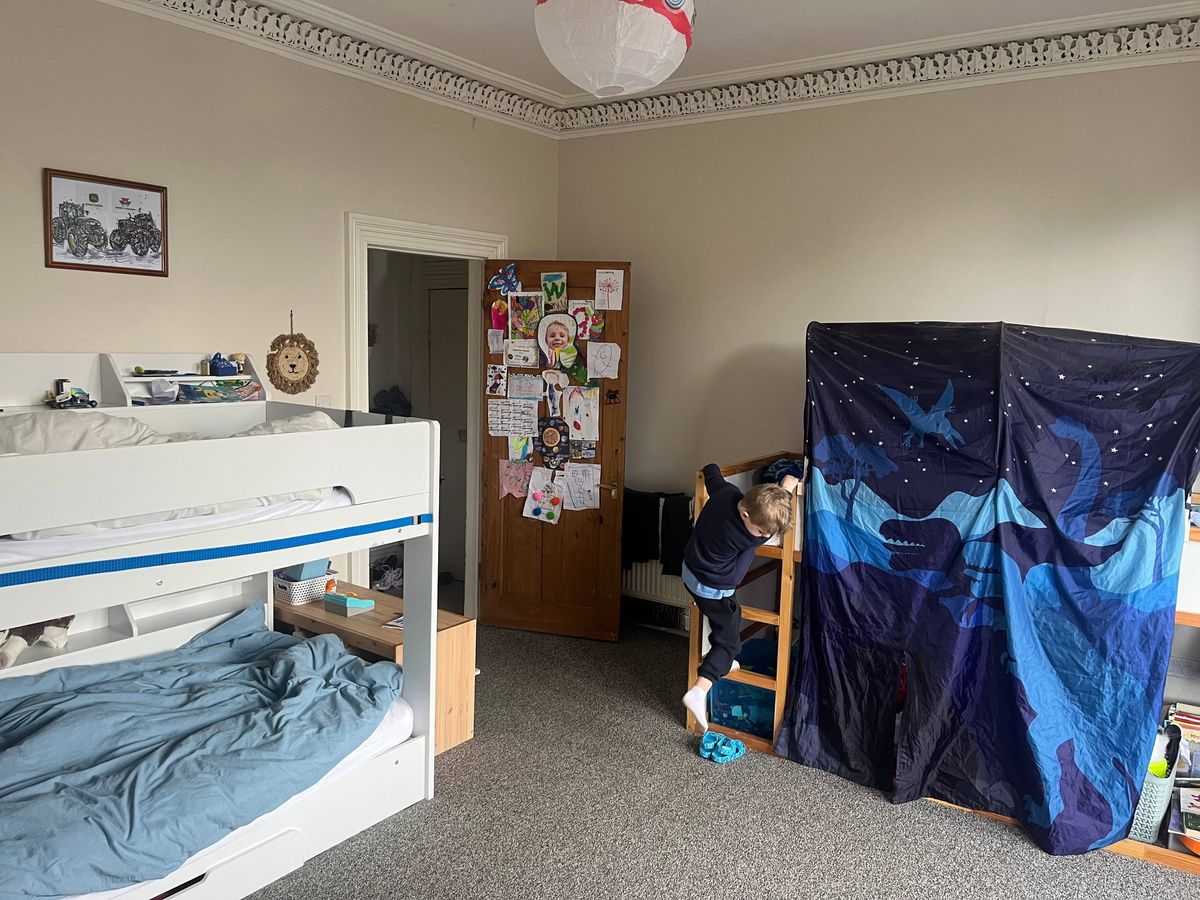 An interior view of a bedroom featuring a white bunk bed, a wooden door, a framed picture on the wall, a blue partition curtain, and various items on a shelf.