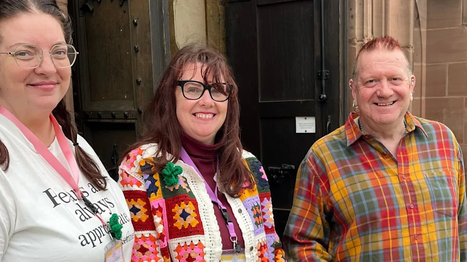 Two women, one with brown hair in pigtails and one with long dark brown hair, and a man with hair dyed red, smile at the camera outside the front door of a church.