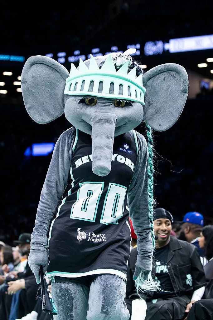 A sports mascot dressed as a stylized elephant with a basketball jersey and a crown-like headpiece at an indoor sports event