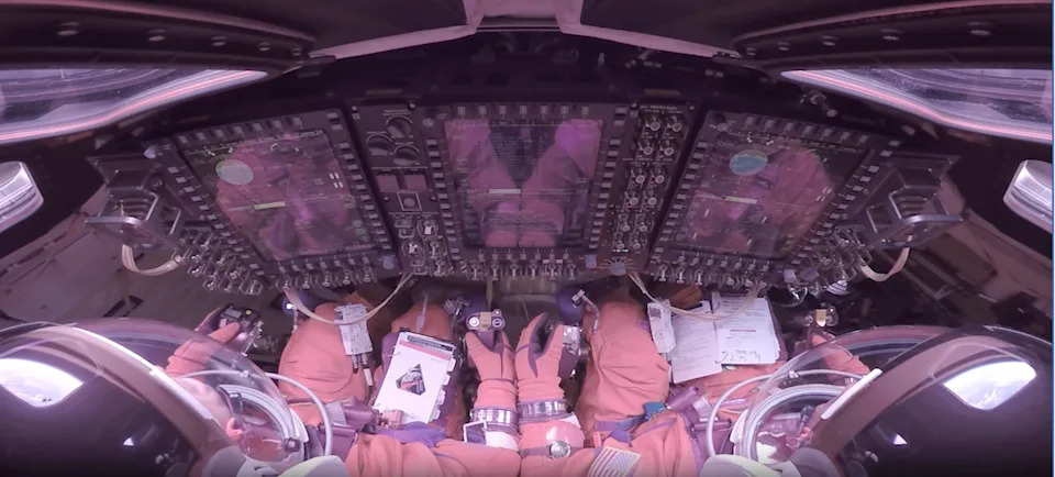 This image looks forward over the shoulders of two Artemis II astronauts strapped into their seats inside the Orion crew module. They wear bulky orange pressure suits and clear helmet bubbles, facing a wall of tall, glowing display screens and dense rows of switches and knobs. Their gloved hands rest near hand controllers and a checklist. Around the edges of the frame, curved windows, cables and equipment make the cabin look compact and highly technical