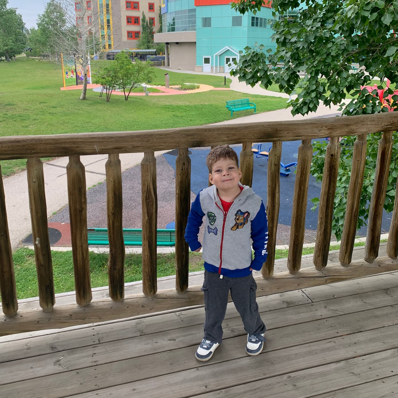 A boy wearing a grey and blue hoodie stands on a deck in front of Alberta Children's Hospital.