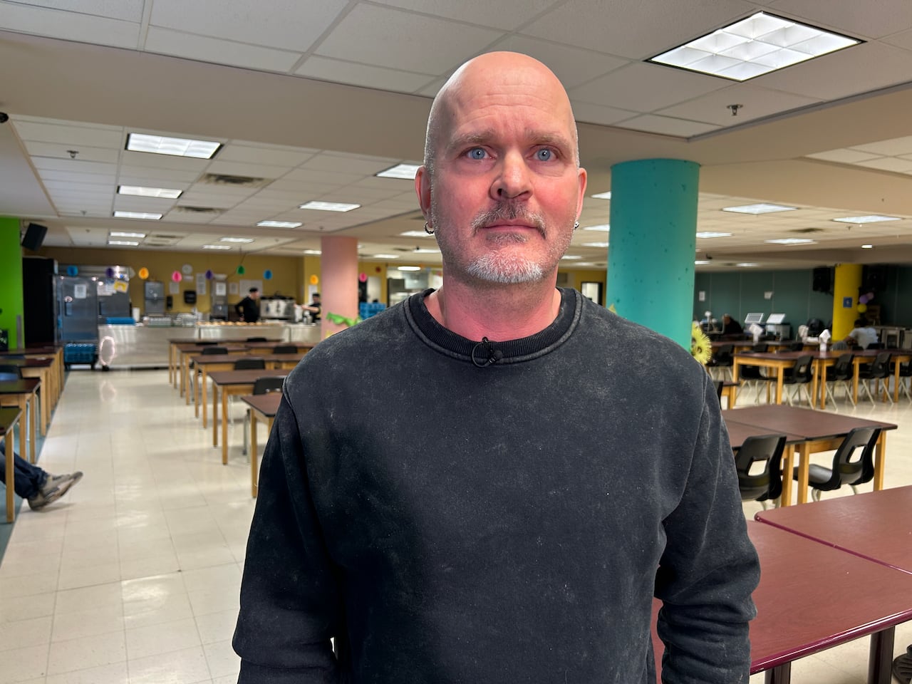 A man in a black sweather poses for a photo inside a cafeteria-style room. 