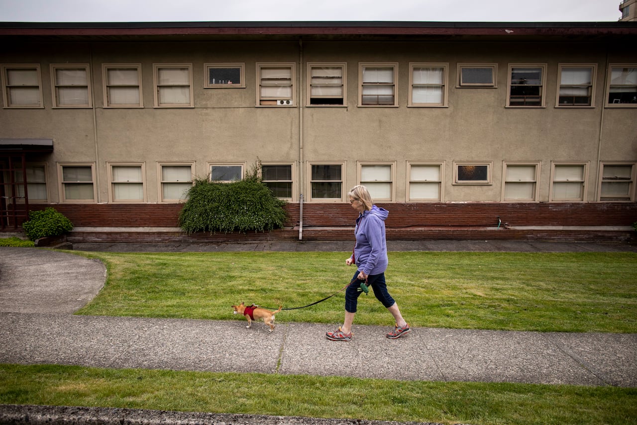 A woman walks a small dog on a sidwalk in front of a beige multi-unit apartment building.