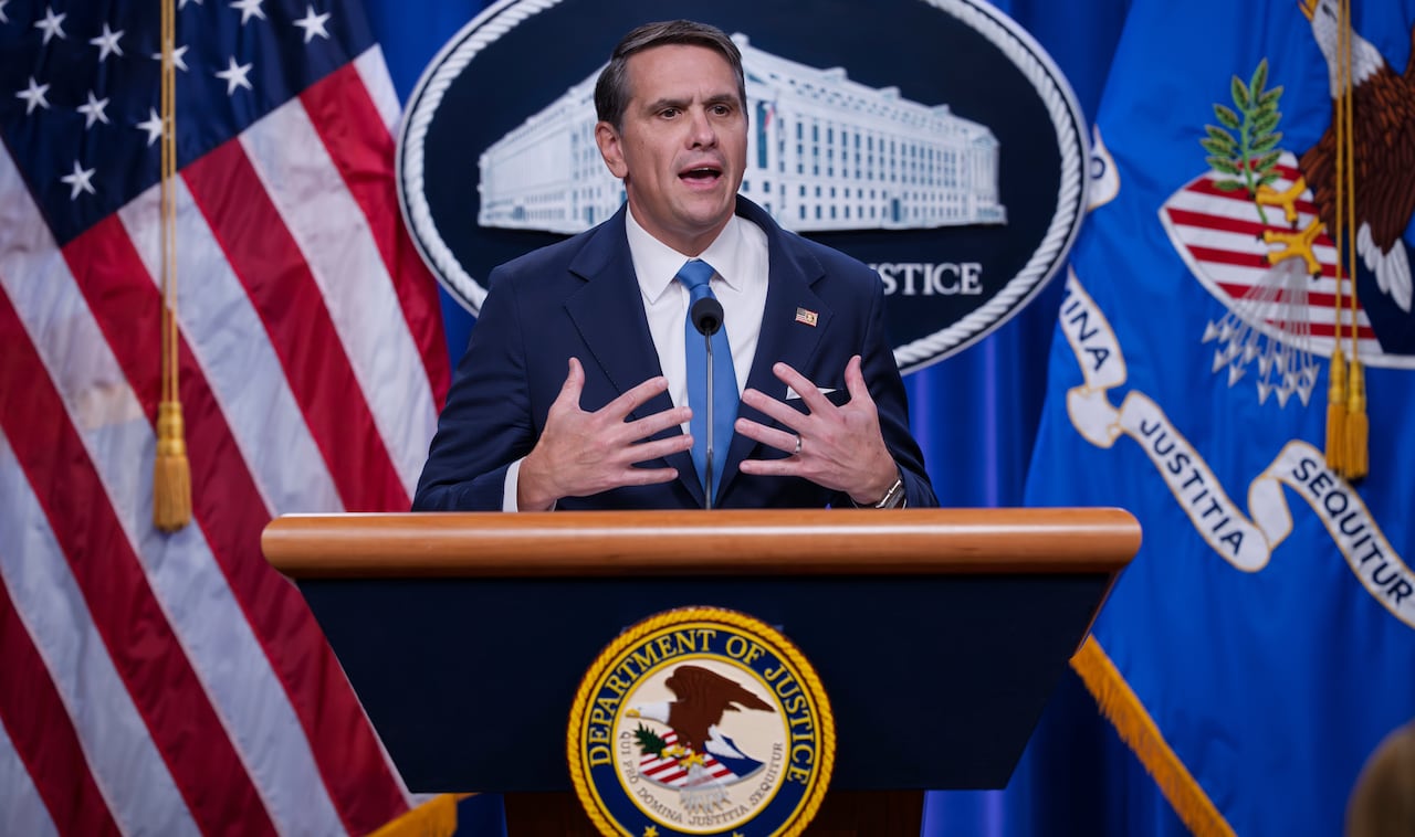 Todd Blanche stands at a podium marked with a Department of Justice seal in front of a backdrop of flags. 