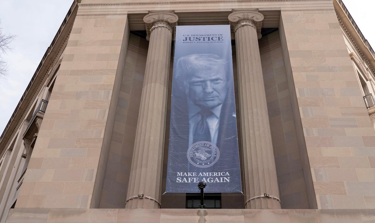 A large banner picturing Trump, with the words 'Department of Justice' and 'Make America Safe Again,' hangs between the columns of a building.