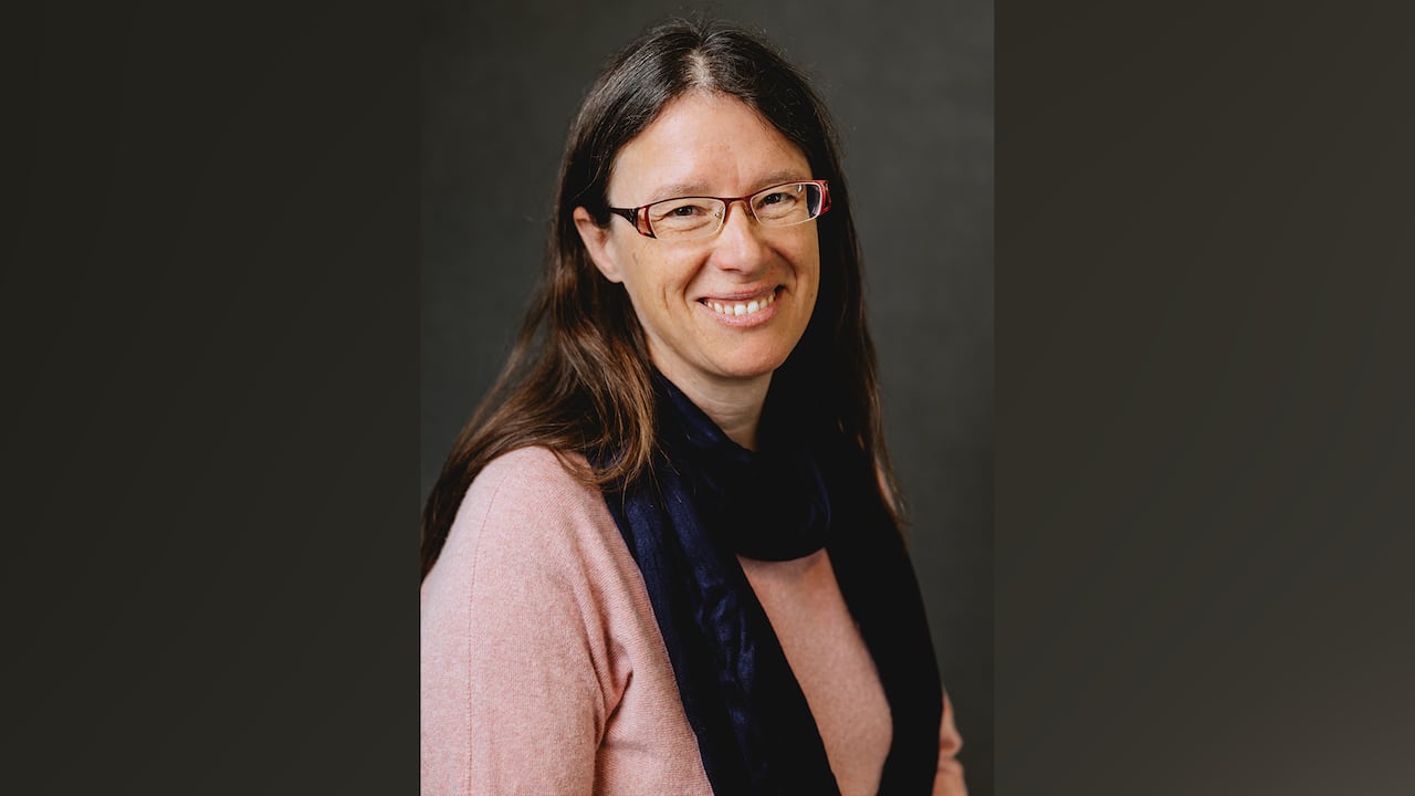 A portrait of a smiling woman with long dark hair, glasses and wearing a dark scarf over a pink sweater.