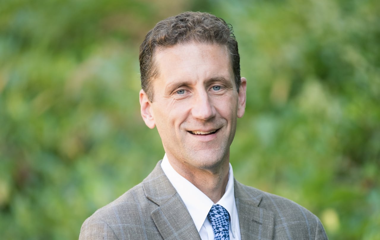 A smiling man in a brown checked suit and blue tie is seen in this outdoor portrait image.