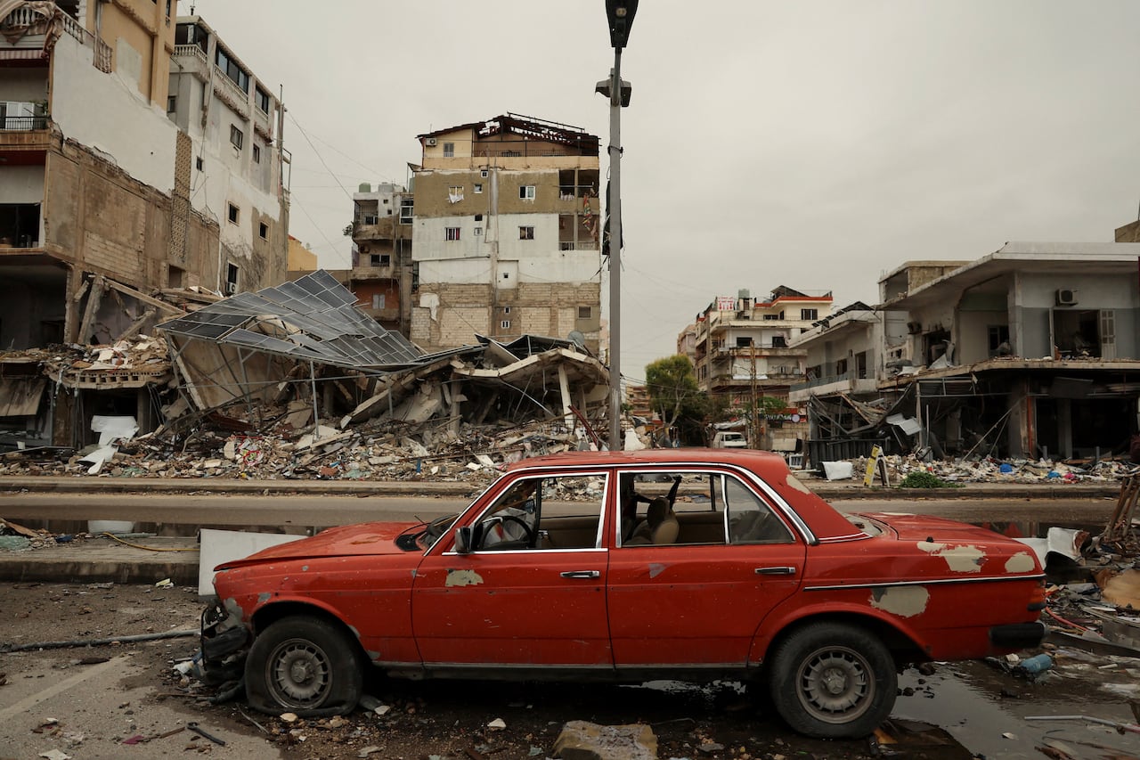 A red car sits on a road strewn with rubble. In the background are damaged and destroyed buildings.