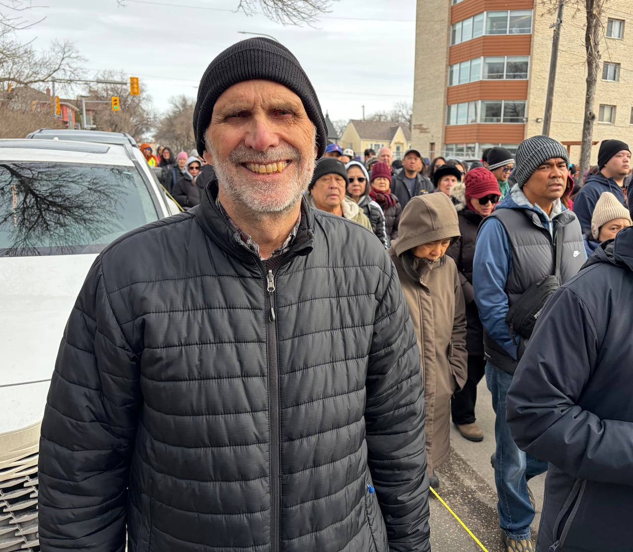 A man in a jacket and toque smiles as a crowd of people walks behind him.
