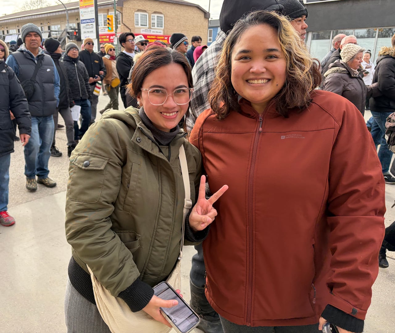 Two women smile in front of a crowd of people. One of them makes a peace sign with her hand.