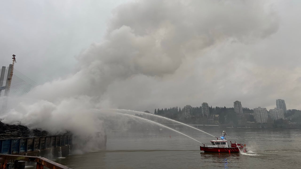 Firefighters on a boat attack a barge that gives off smoke into the air on the water