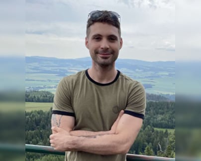 A man leans against a railing and smiles, with arms crossed in front of him. Rolling farmland and forest can be seen in the background.