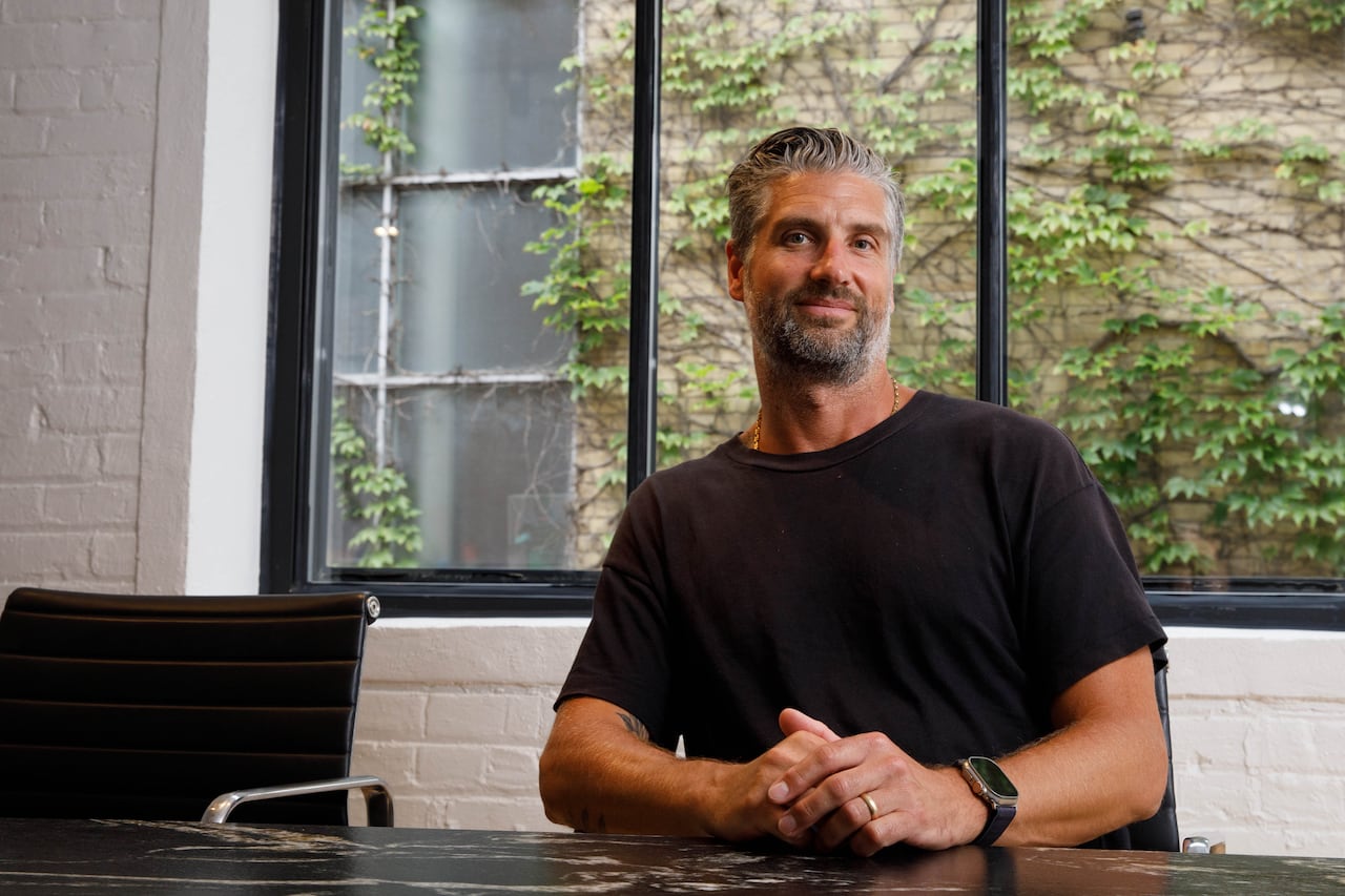 A man in a black T-shirt sits at a desk and poses for a photo. Behind him is a large window.