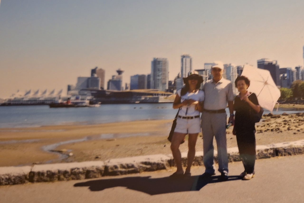 Three people stand near a beach, with Vancouver's downtown in the background.