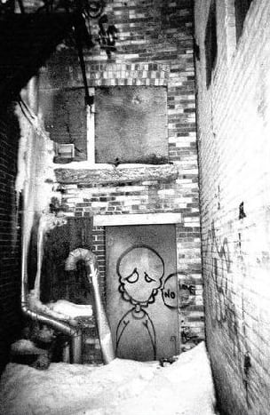 A spray-painted character on a door in a snowy alley.
