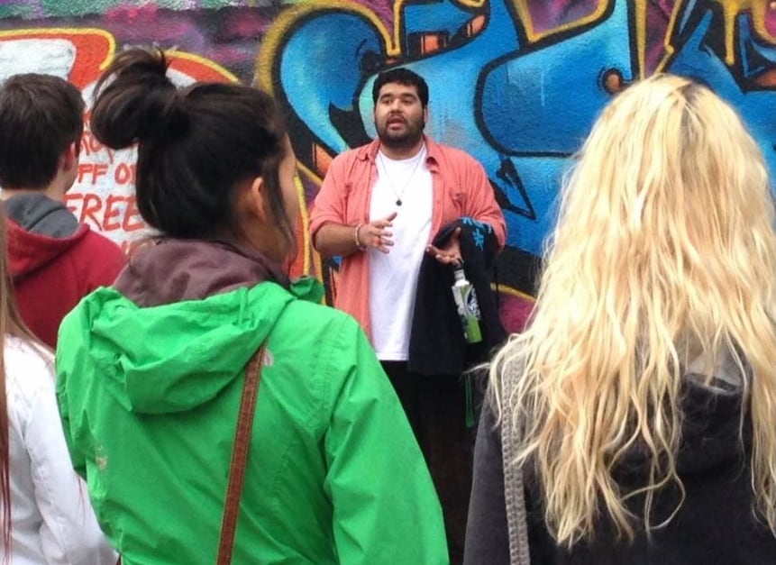 A man speaks in front of a wall painted with colourful murals.