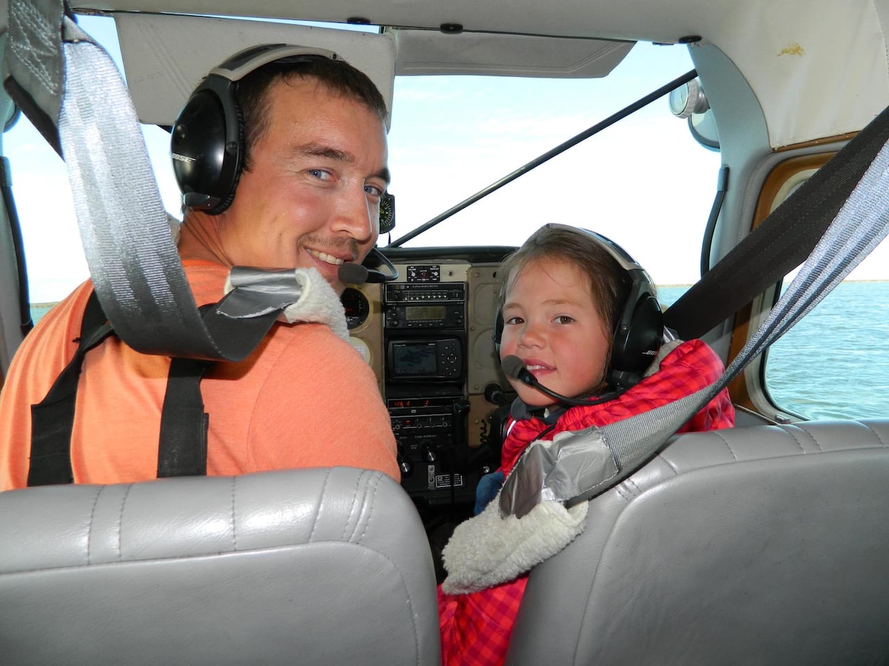 A man in an orange shirt and pilot's headset and a child in a pilot's headset look to the back of a small plane from the cockpit and smile.