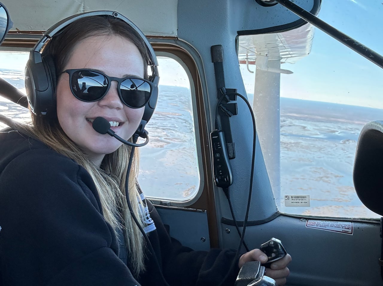 A smiling young woman with long hair wears sunglasses and a pilot's headset as she sits in the cockpit of a small plane.