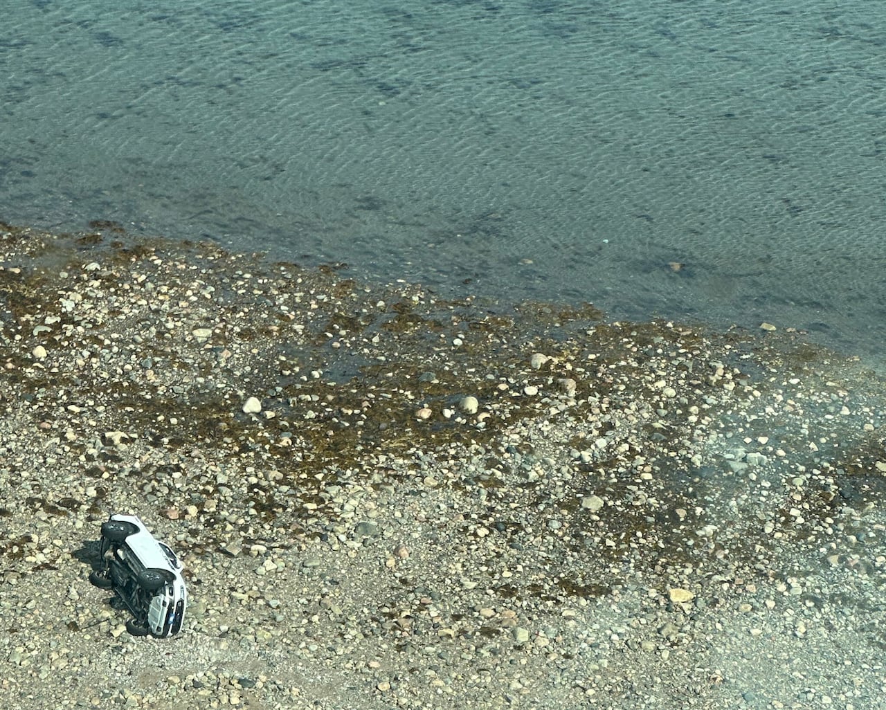 A white truck on its side in the rocky shore near a large body of water