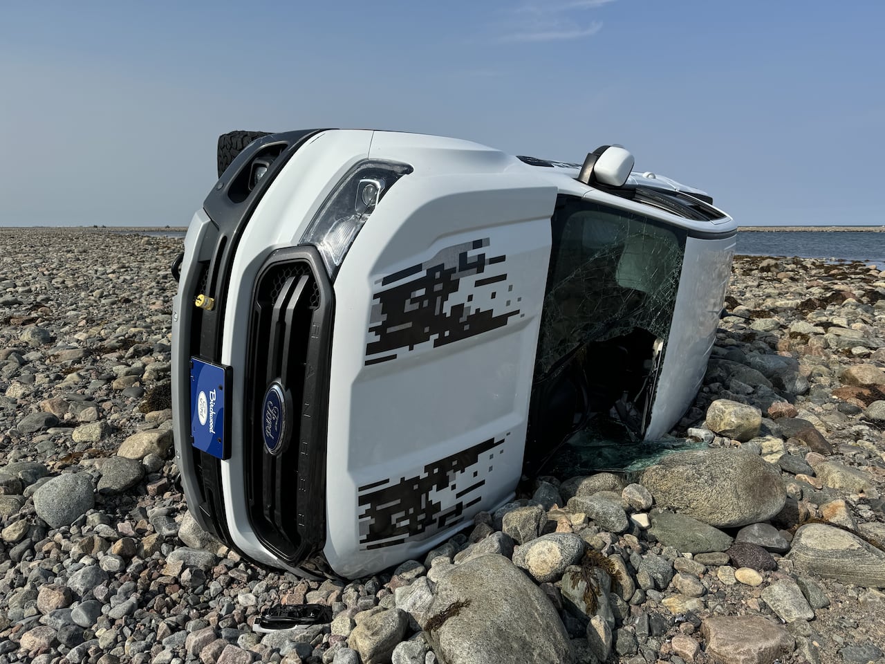 A white truck lies on its side on a rocky shoreline.