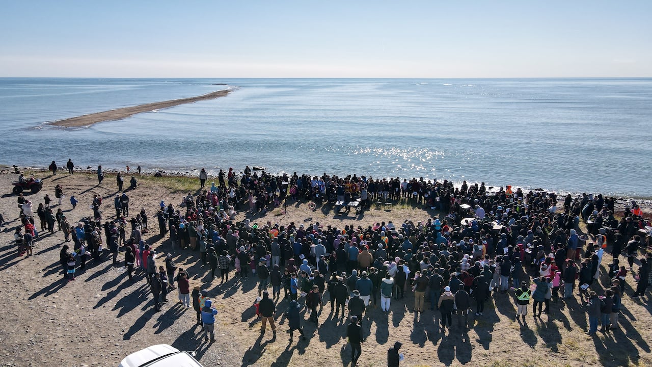 A drone photo shows a large group of people forming a circle around a family on the shoreline next to a large body of water. A strip of land emerging from the water can be seen on the left.
