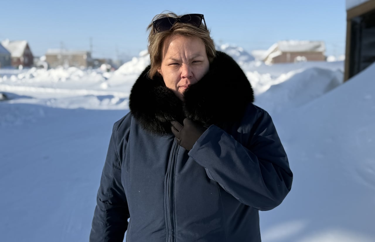 A woman with short, red hair wears sunglasses on her head and a dark blue parka with black fur trim as she stands in the snow and squints into the sun.