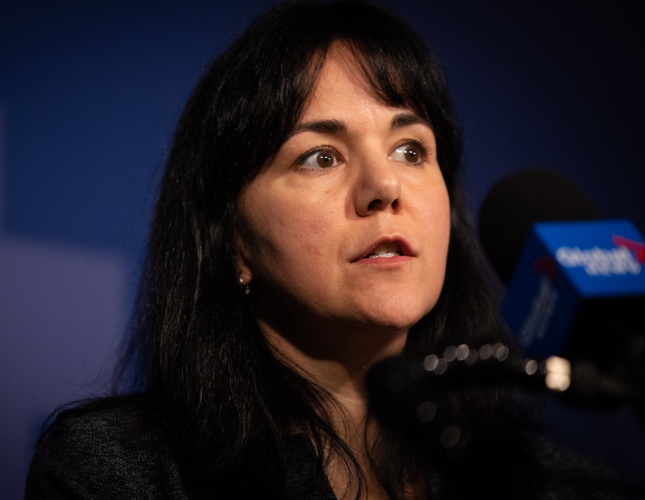 A woman with black hair speaks to a number of mics.
