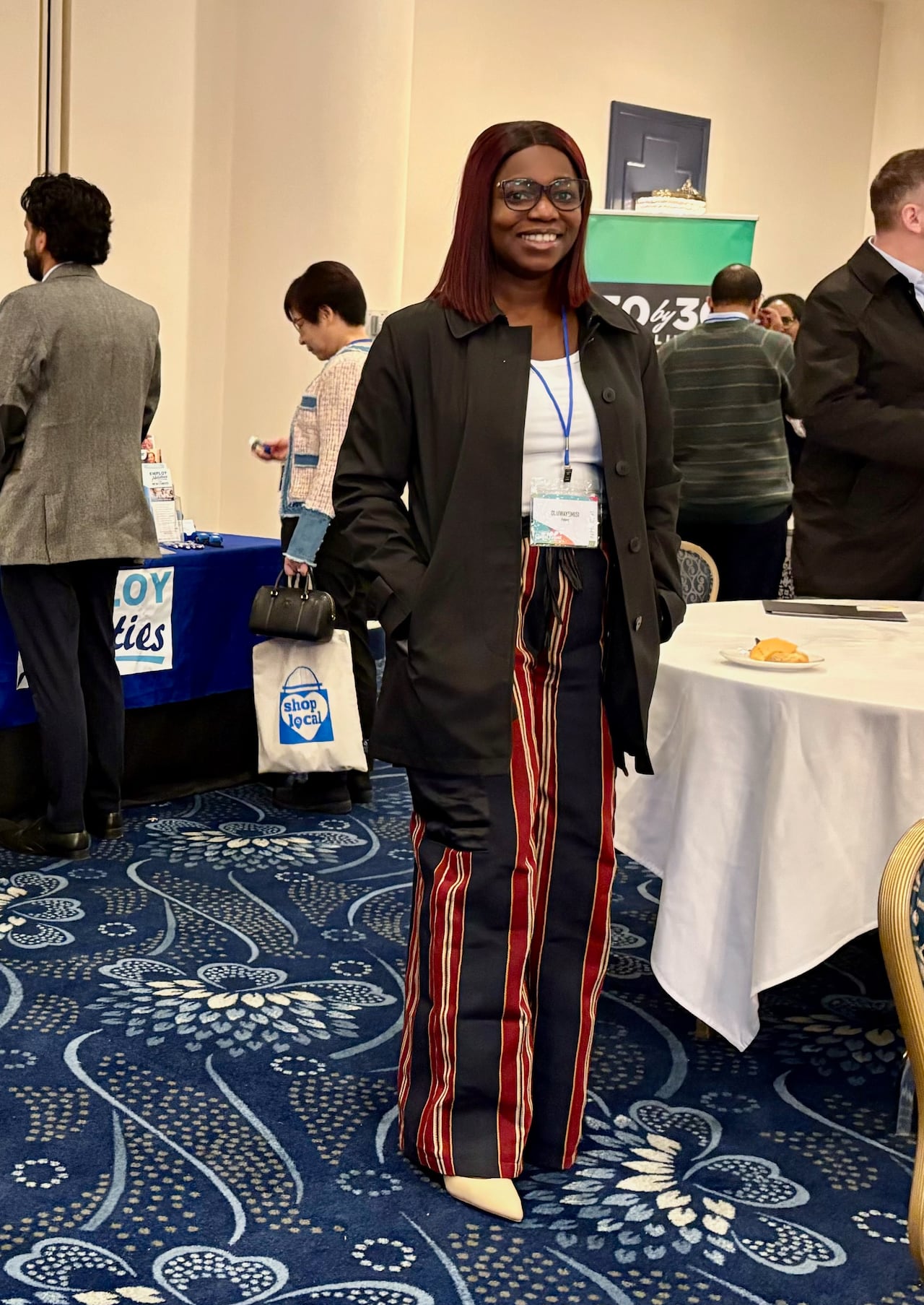 A smiling woman wearing red and blue pants poses for a photo in a conference room. Other attendees are milling around.