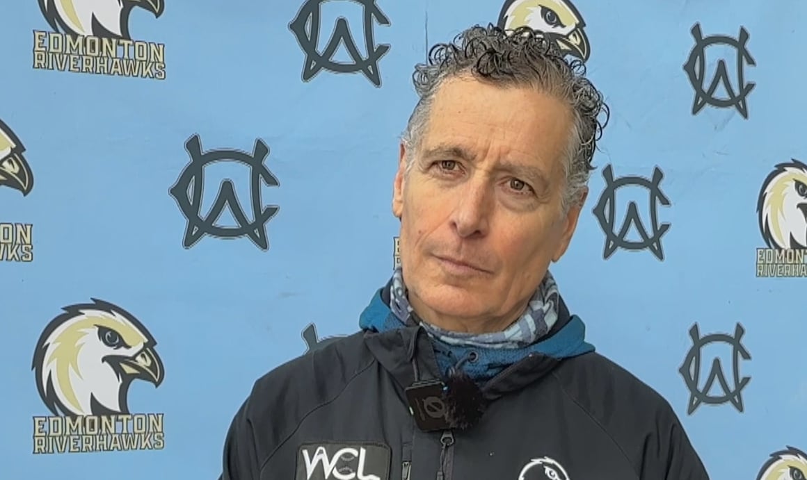 A middle-aged white man, with short grey hair, is wearing a navy blue windbreaker while standing in front of a light-blue screen that features logos hawks