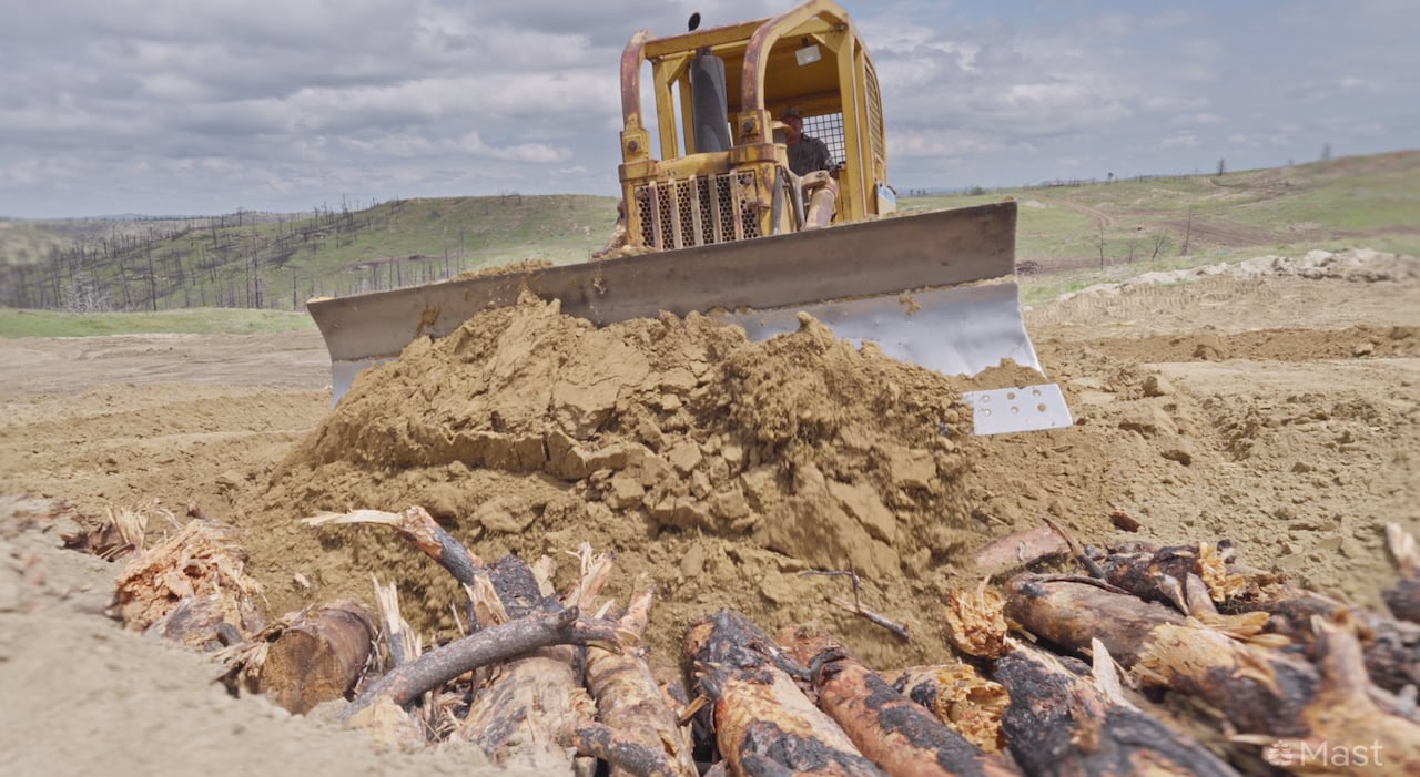 A yellow bulldozer pushes dirt over burnt logs on the ground.