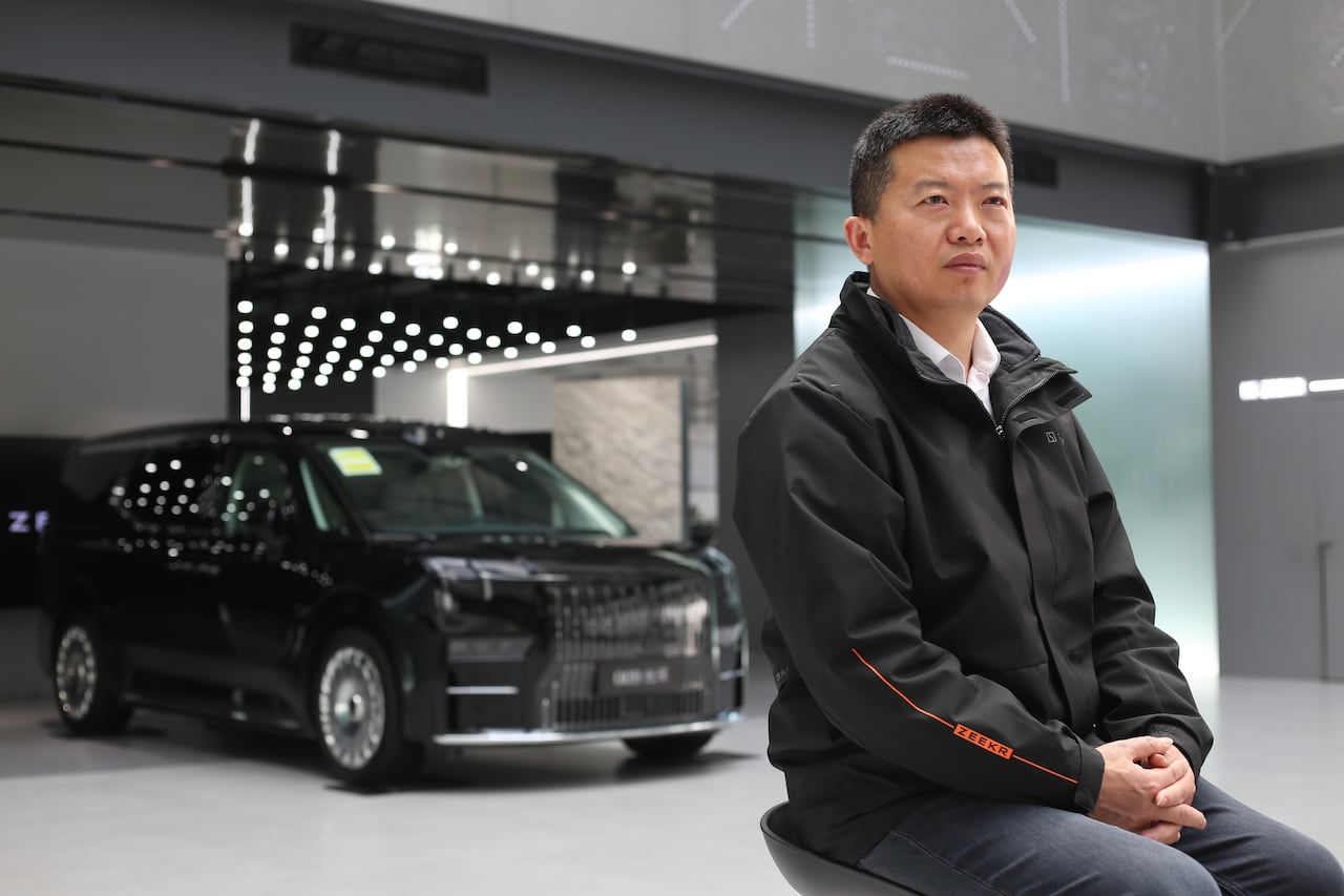 A man in a black jacket sits at right, with a black car behind him