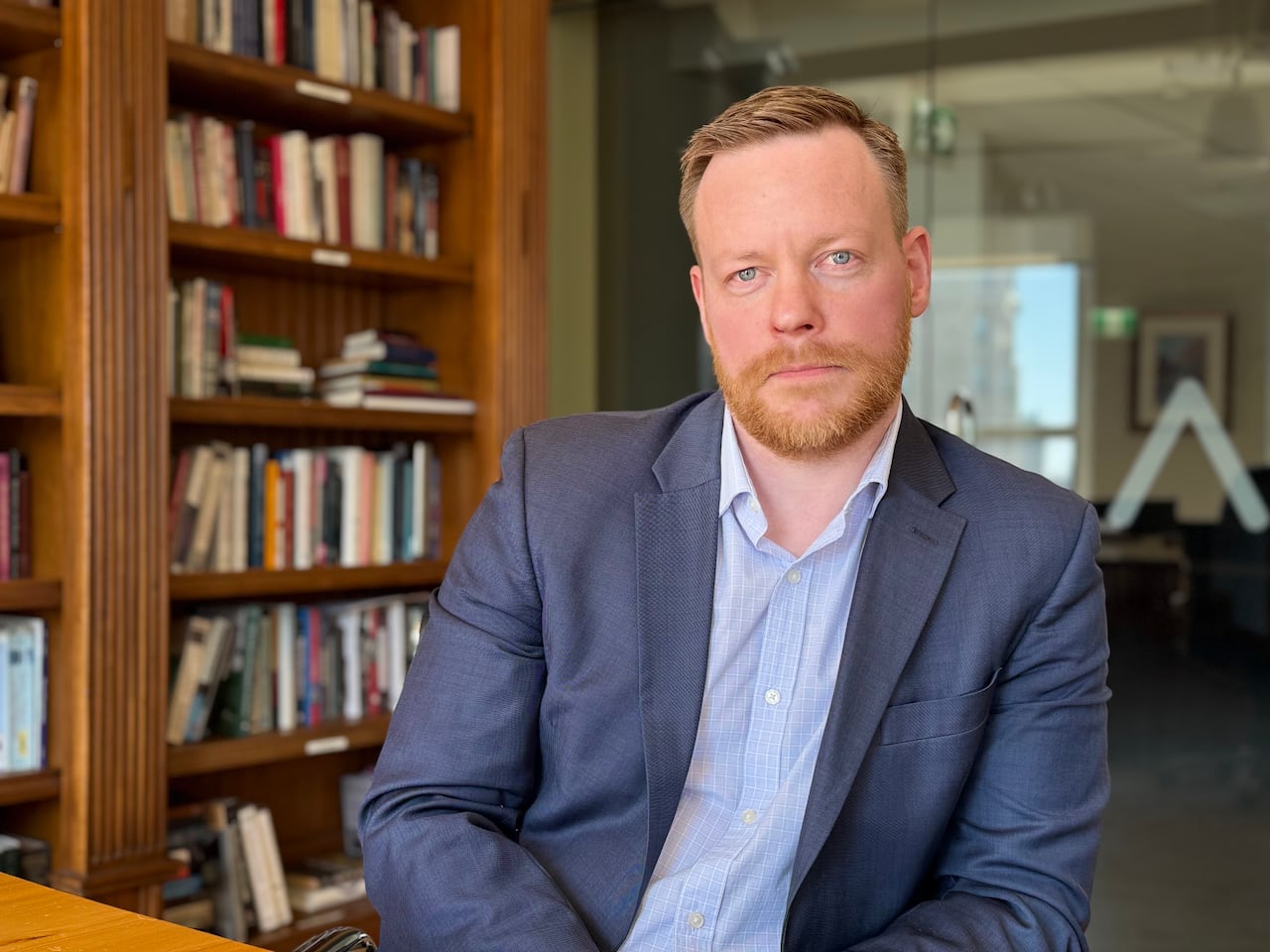 A man with red hair and a beard wears a suit in front of a book case. 