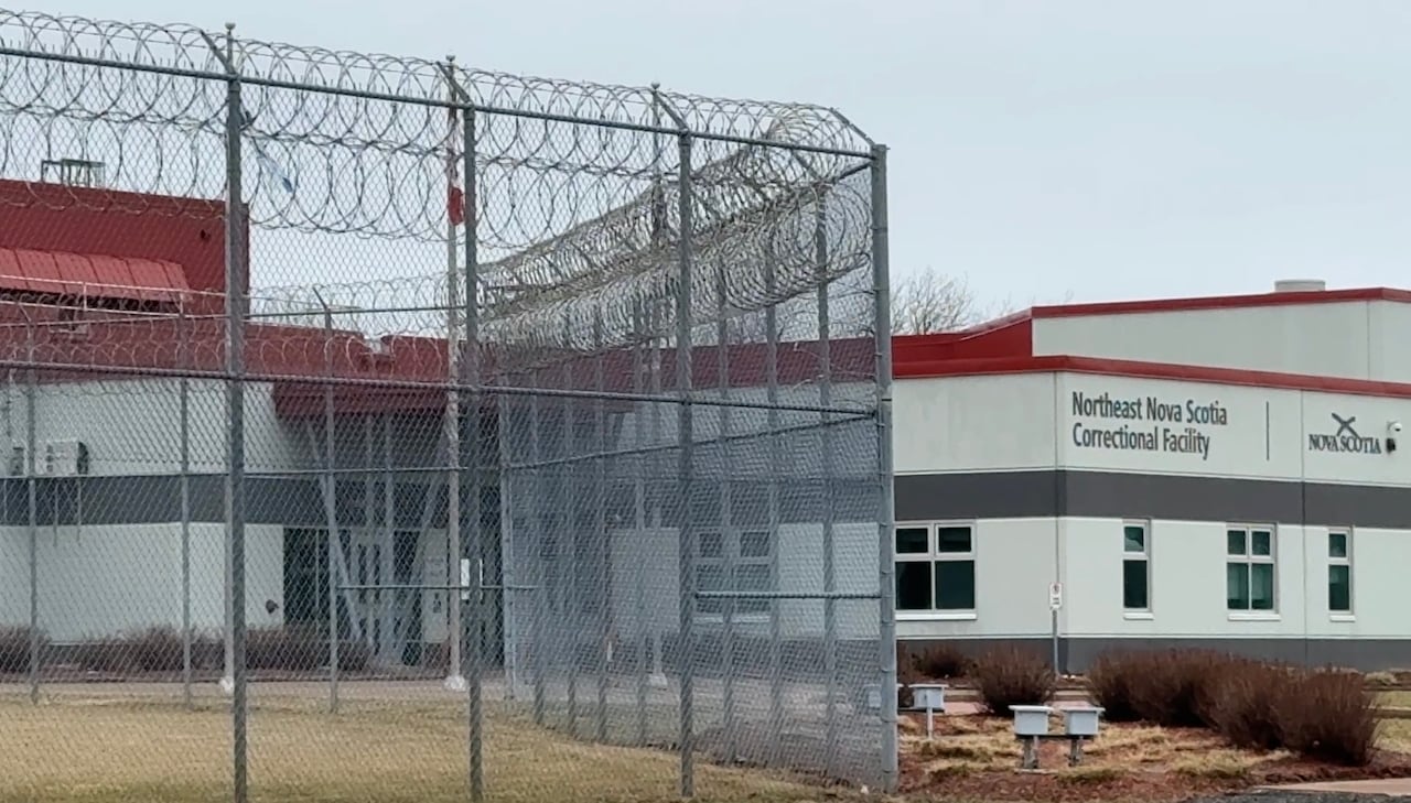 A tall fence with barbed wire and a building in the background is shown.