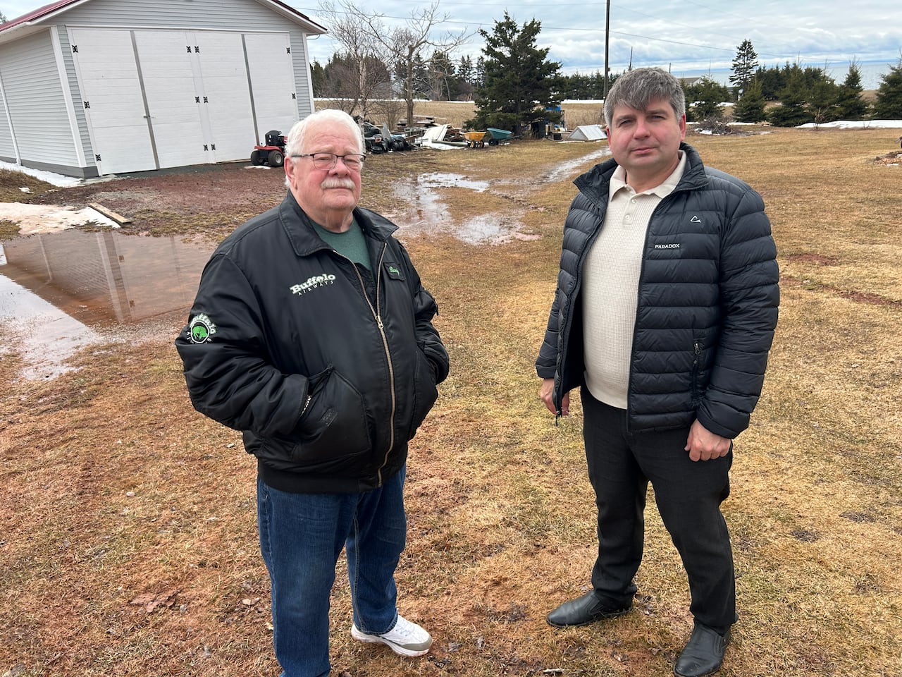 Two men stand outside in a yard with water pooling on the lawn behind them. 