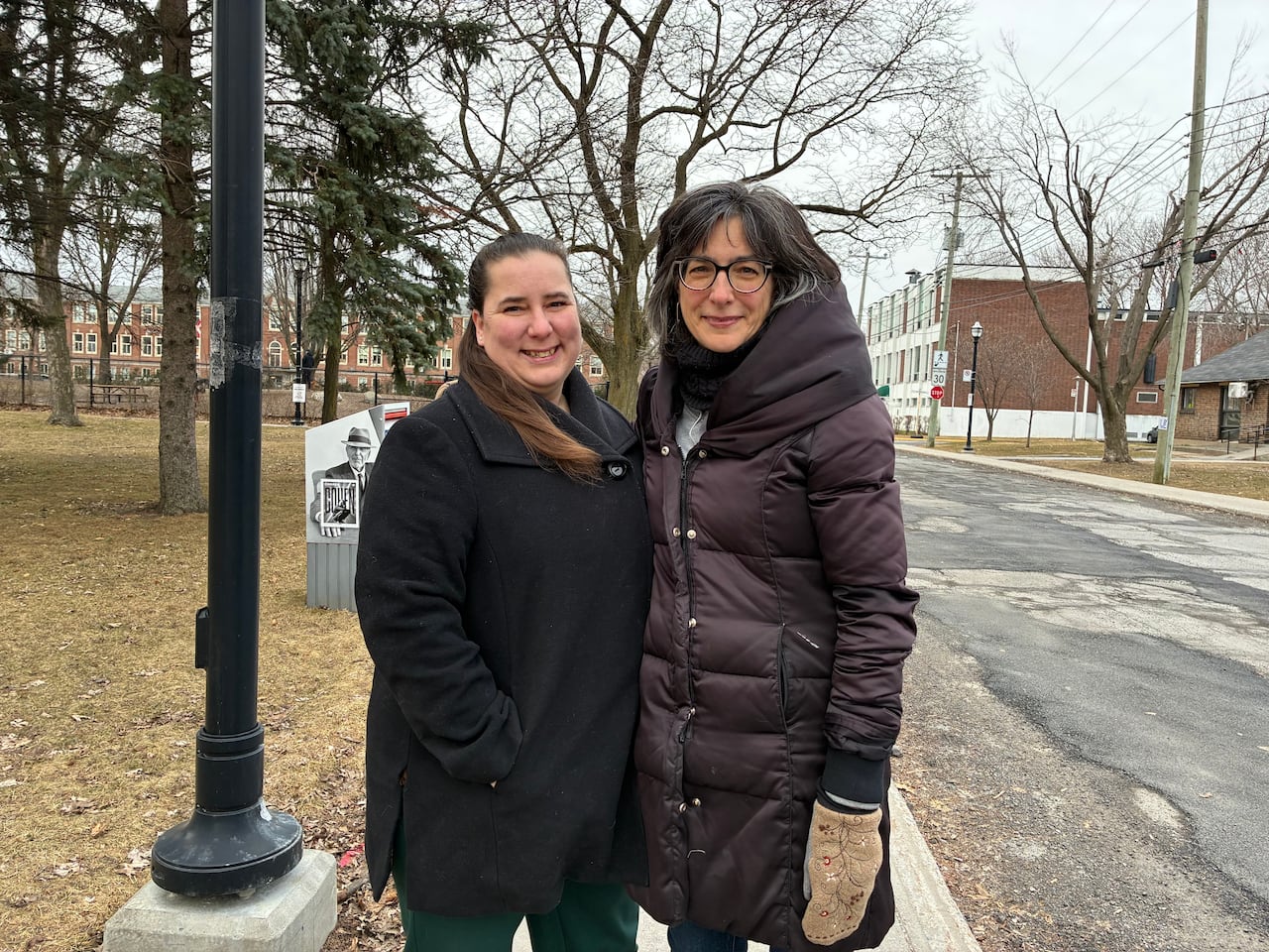 Two women stand on a sidewalk with a brick building in the background across the street.