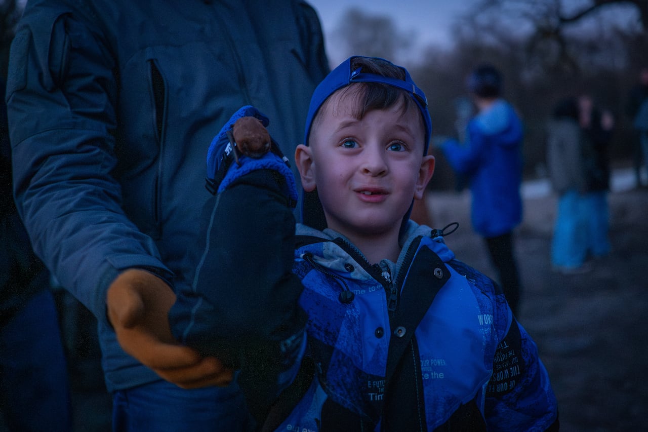 A little boy at dusk, standing next to an adult, grins joyfully as he stares into the sky 